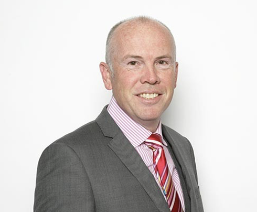 Man in gray suit, pink dress shirt, and red striped tie smiling against a white background.