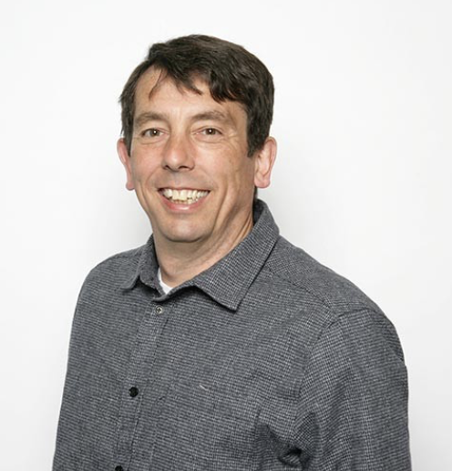 A smiling man with dark hair, wearing a gray button-up shirt, against a plain white background.