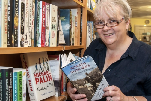 A smiling woman with glasses and short blonde hair holding a book titled 'Beneath Safer Skies' in a bookstore or library, standing next to a bookshelf filled with various books.