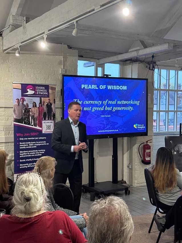 A man in a suit giving a presentation in front of an audience in a room with white brick walls and large windows. A large display screen behind him shows the title "PEARL OF WISDOM" with a quote about networking. There is a banner on the left side promoting the Severn Business Network.