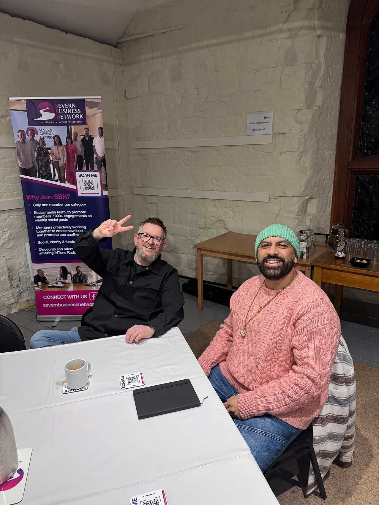 Two men sitting at a table, smiling and posing for the camera. One is wearing a black shirt and glasses, the other is wearing a pink sweater, blue jeans, and a green beanie. There is a poster in the background for Severn Business Network and a mug on the table.