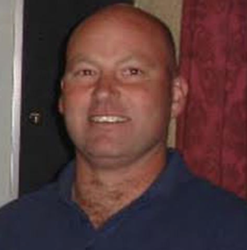 A man with a shaved head wearing a navy blue shirt, smiling, standing indoors with a red curtain in the background.