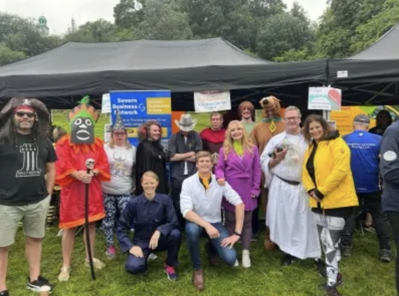 Group of people standing and sitting outdoors under a black canopy tent at an event, with some dressed in costumes and casual clothing, with trees in the background.