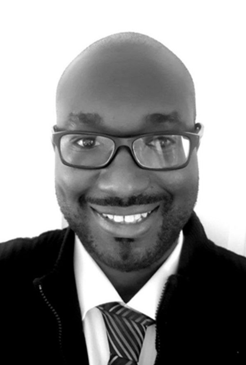 A smiling man with glasses, wearing a suit with a tie, in a black and white photo.
