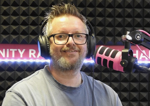 A man with glasses, a beard, and spiky hair wearing headphones, speaking into a pink and black microphone in a recording studio.