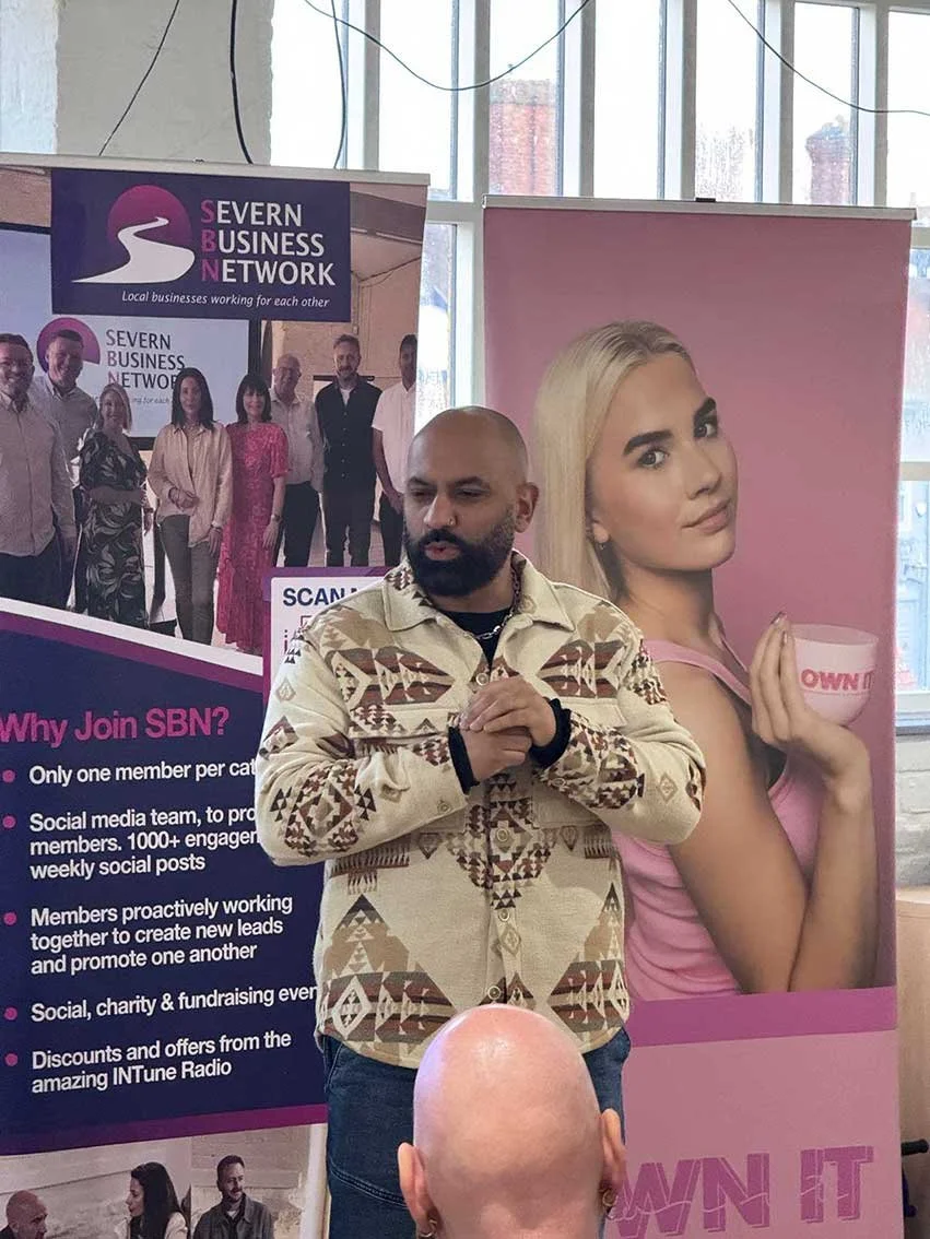A man with a beard and shaved head wearing a beige patterned jacket standing in front of two banners, one with a group photo for Severn Business Network and the other with a large image of a woman with blonde hair, pink top, holding a bowl. The man is holding his hands together and looking down, with part of a bald person's head visible in the foreground.