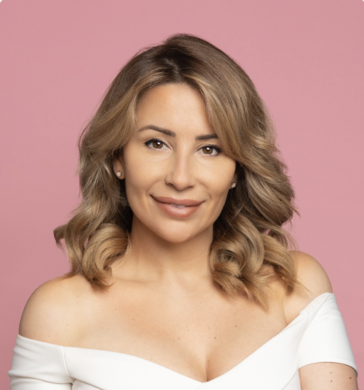 A woman with shoulder-length wavy blonde hair, smiling, wearing a white off-shoulder top, against a pink background.