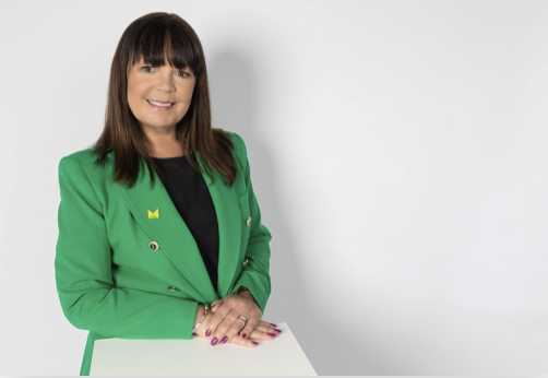 A woman with shoulder-length dark brown hair, wearing a green blazer and a black top, smiling and standing against a plain white background.