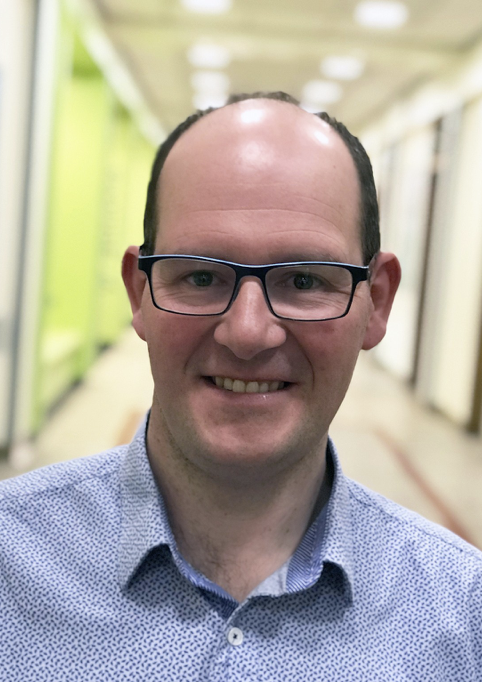 A man wearing glasses and a light blue patterned shirt smiling, standing in a hallway with green and white walls and bright overhead lighting.