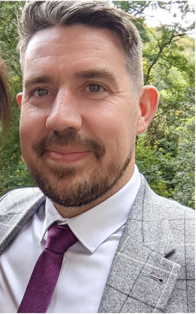 A man with brown hair, a beard, wearing a gray suit jacket, white shirt, and purple tie, posing outdoors with trees in the background.
