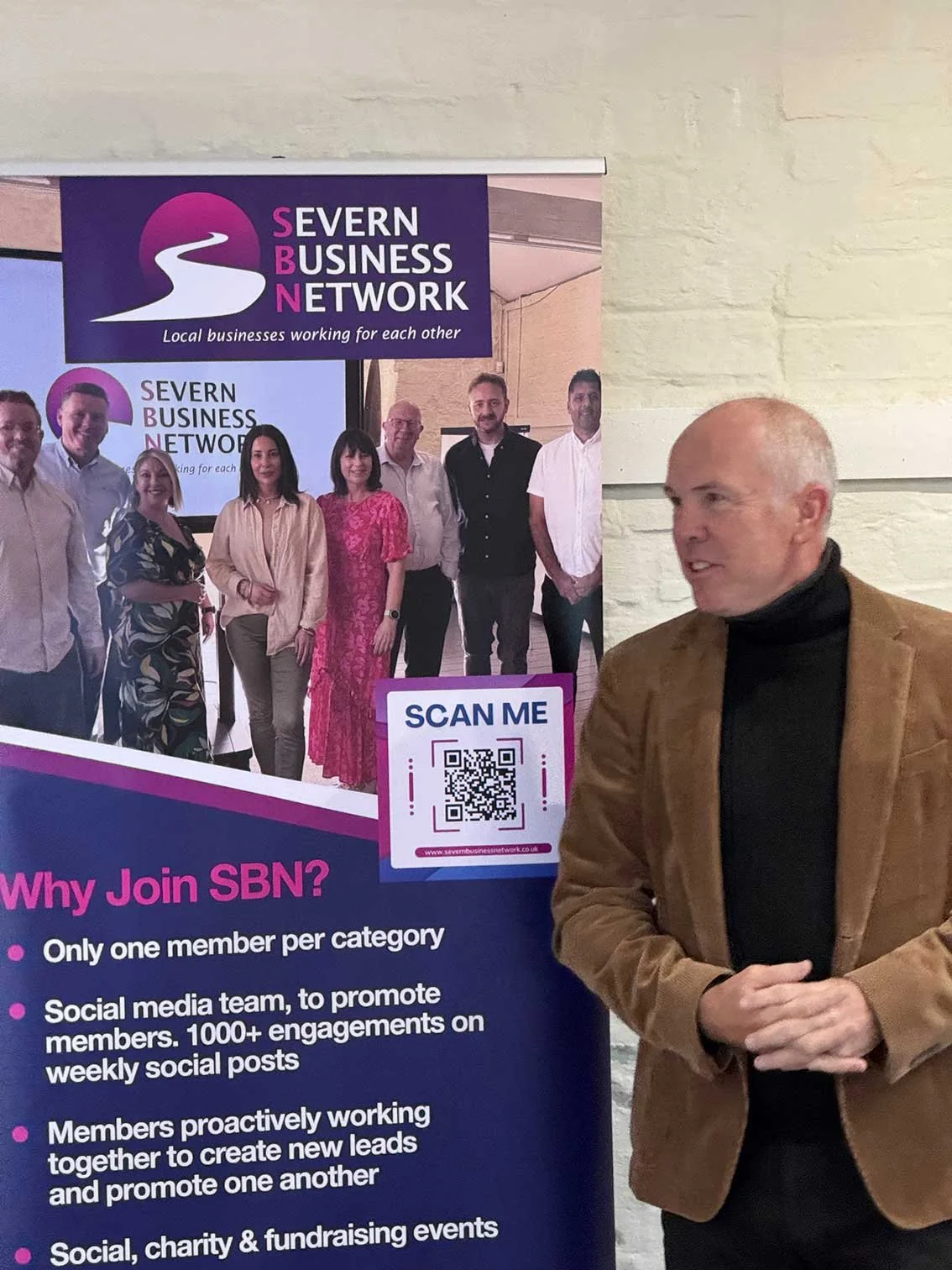 A man in a brown blazer and black turtleneck standing next to a promotional poster for Severn Business Network. The poster features a group photo of people and information about the networking group, along with a QR code and website link.