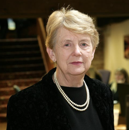 An older woman with short blonde hair, wearing a black top, a black jacket, and a pearl necklace, standing indoors.