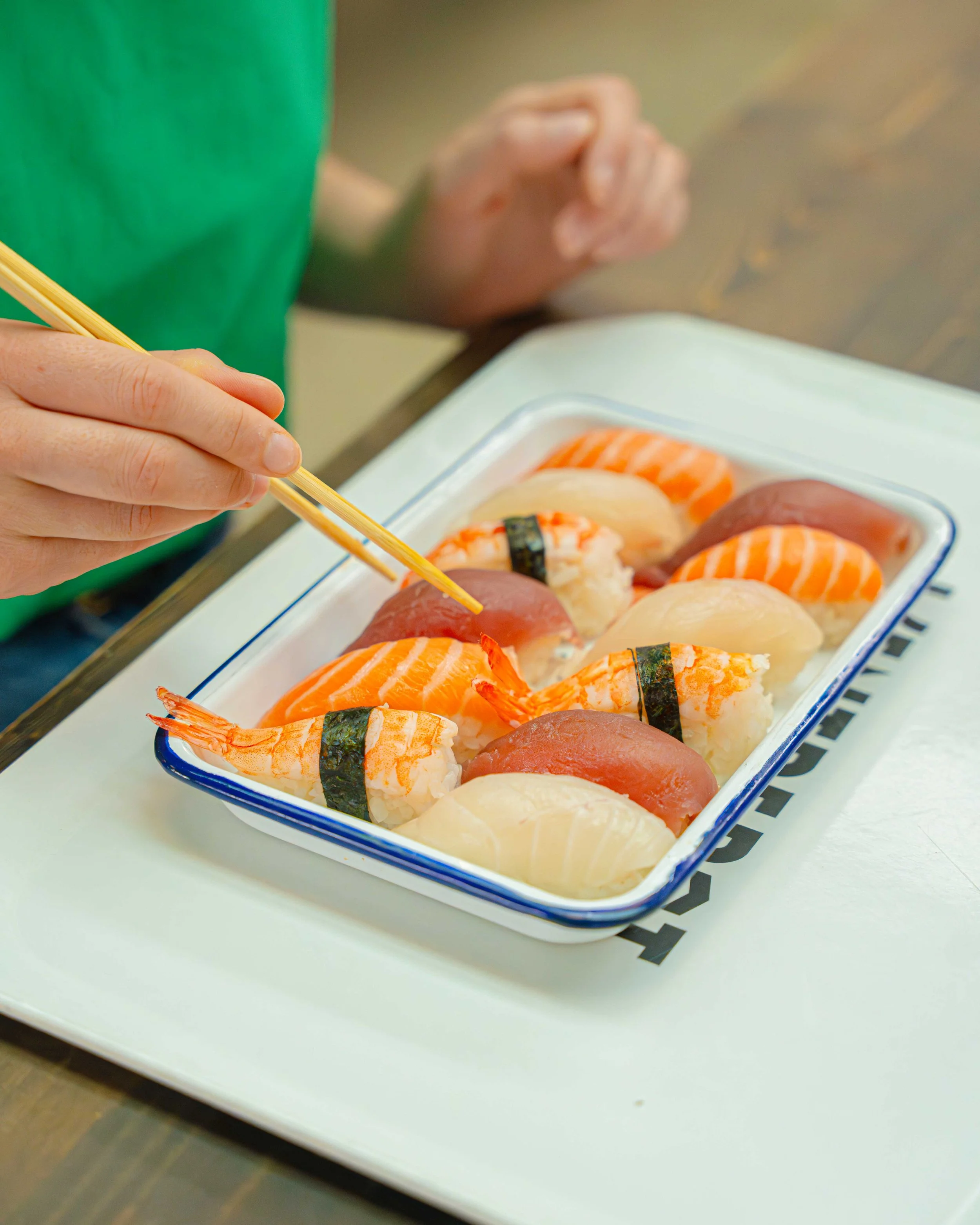 Une personne utilisant des baguettes pour prendre du sushi dans une boîte en céramique, contenant différents types de sushi, sur une assiette blanche.