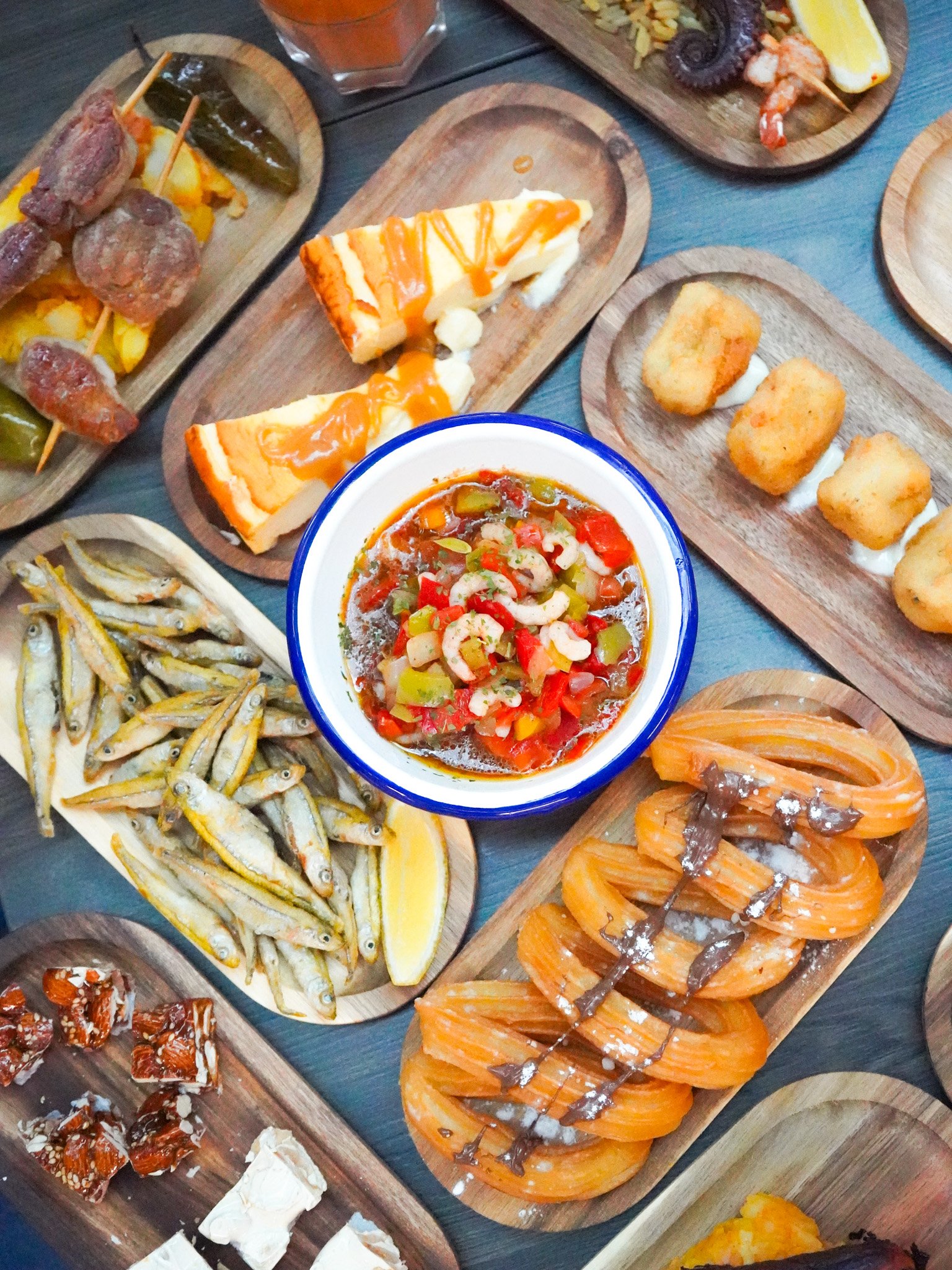Plats de tapas espagnols, y compris des calamars frits, des croquettes, une paella avec des fruits de mer, des sardines, une tartiflette, une salade de crevettes, et des brochettes de viande, tous servis sur des planches en bois.