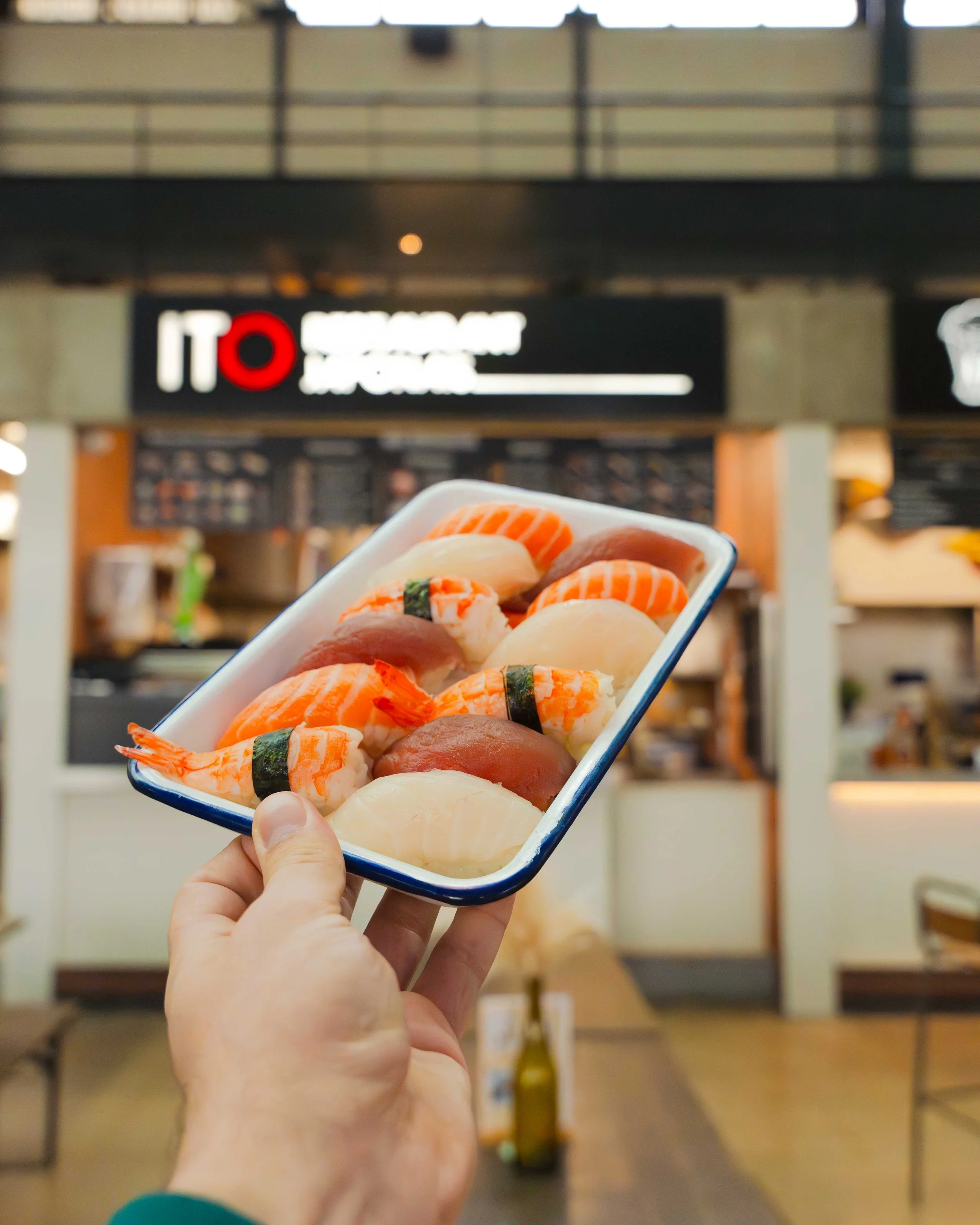 Une main tient une assiette de sushi composée de plusieurs morceaux de poisson cru, crevettes, et fruits de mer, dans un restaurant japonais.