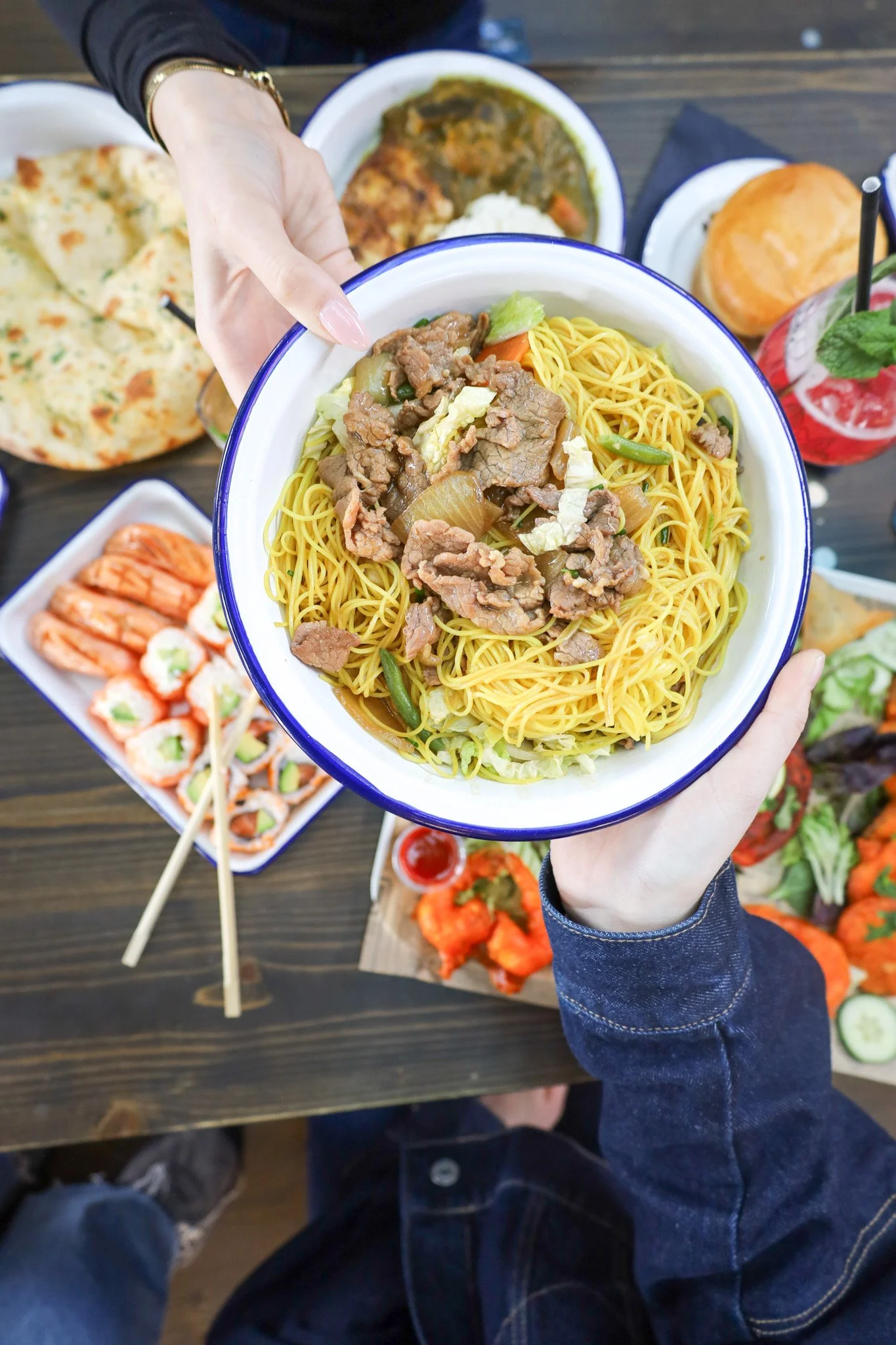 Une personne tenant un bol de nouilles sautées avec de la viande, des légumes verts et de l'oignon. Autour, il y a des plats de crevettes, une pizza, une boisson rouge à la menthe, et d'autres plats variés sur une table en bois.