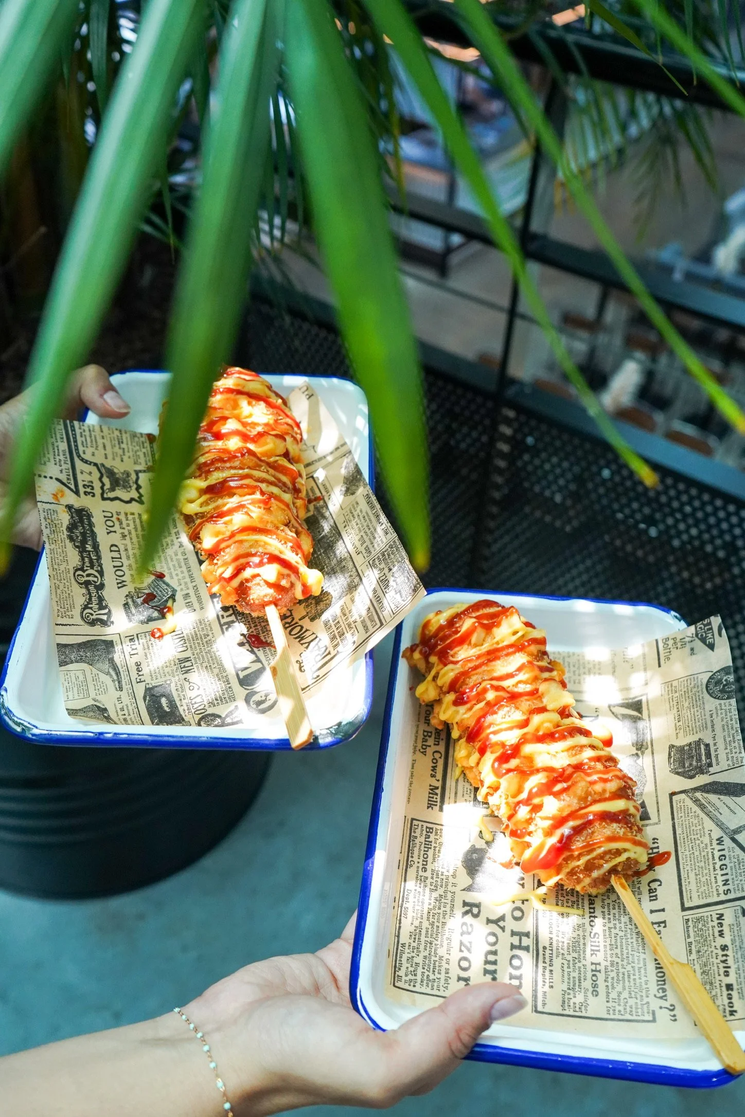 Deux personnes tiennent des assiettes avec des brochettes de fromage enrobé de sauces, posées sur du papier journal. L'une des personnes porte une bague à la main gauche.