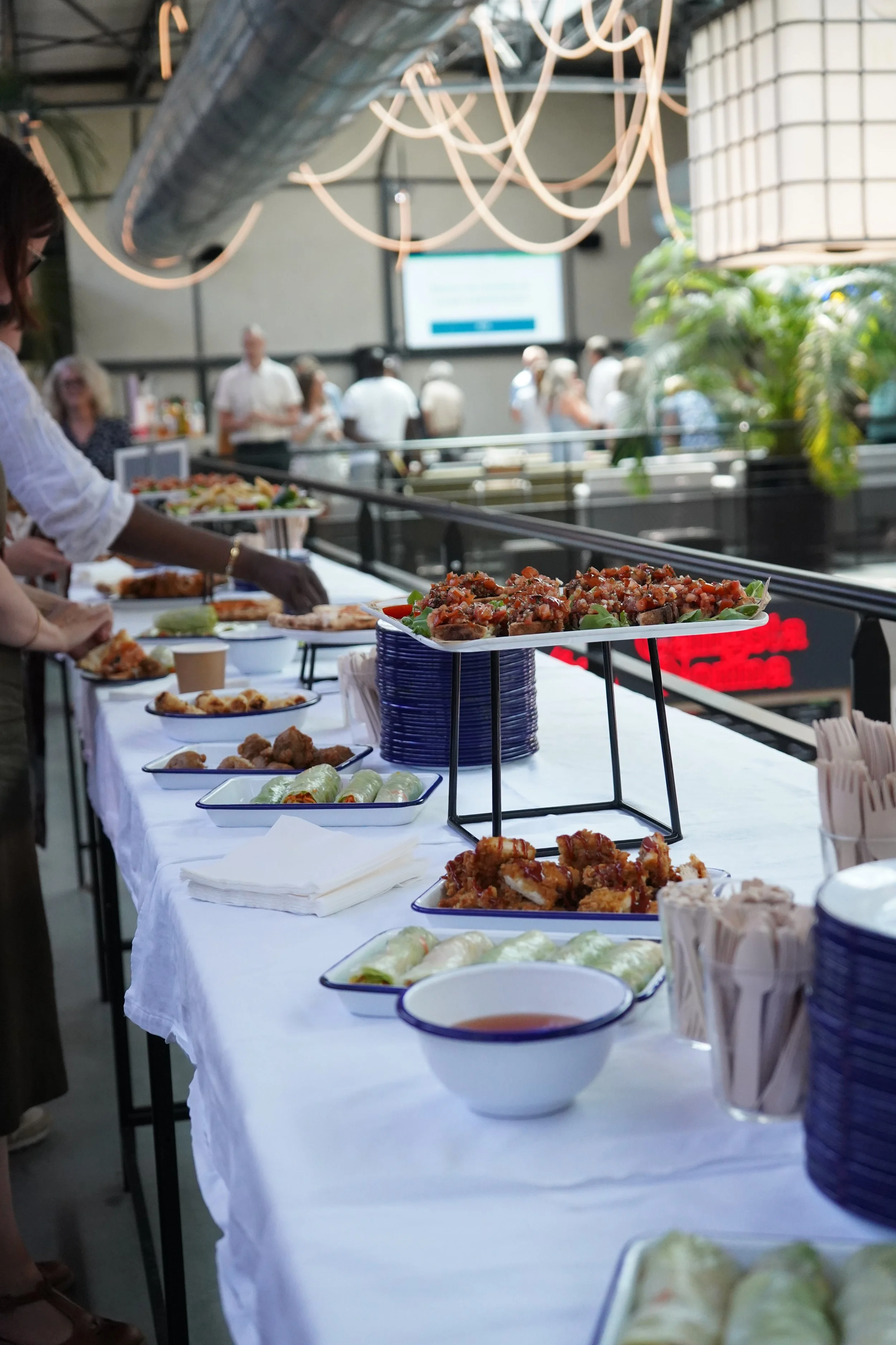 Buffet avec diverses plats asiatiques, notamment des rouleaux de printemps, du poulet frit, et une salade de viande, disposés sur un long table avec une nappe blanche, dans un lieu moderne avec des personnes en arrière-plan.