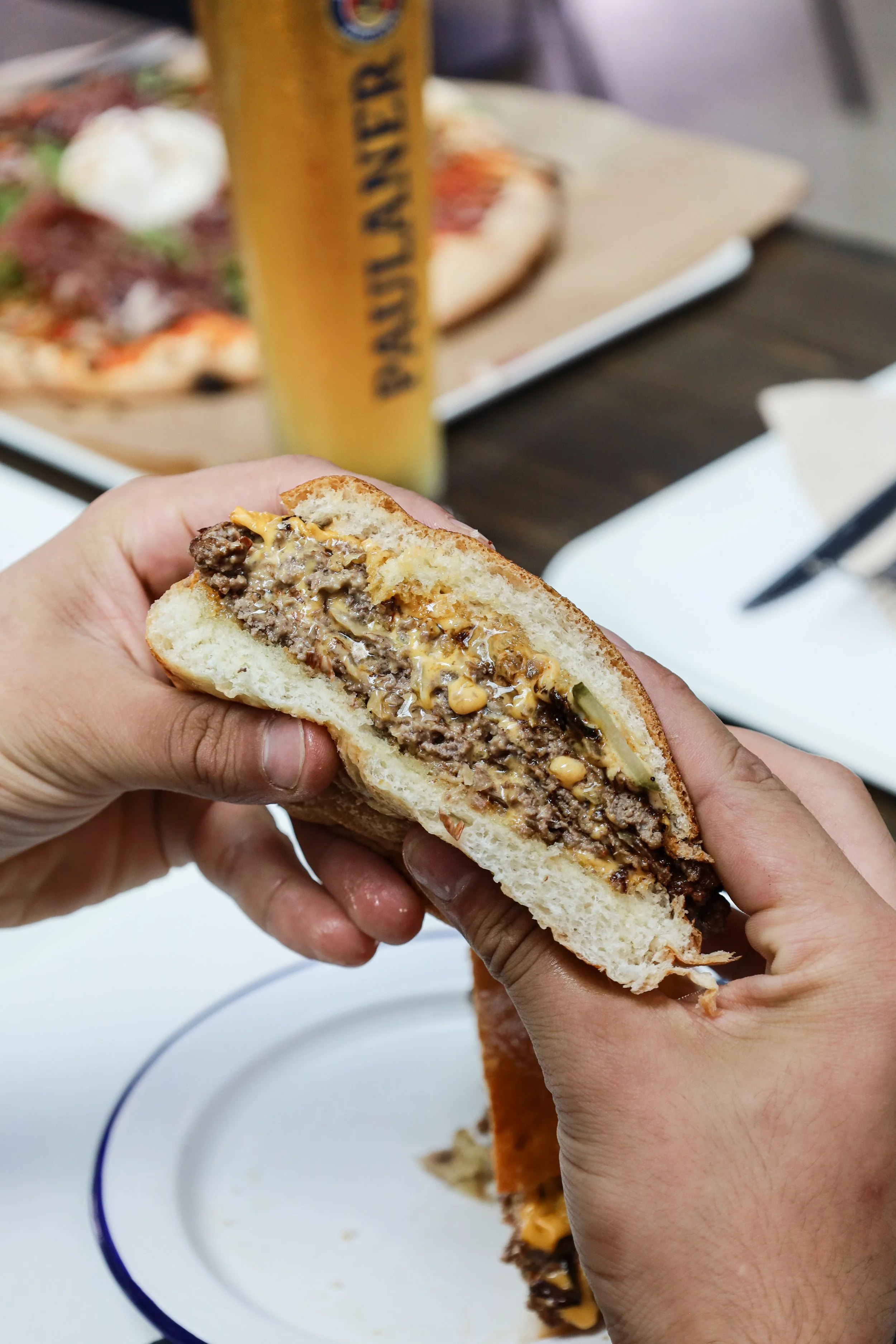 Personnes tenant un burger, avec un fond flou d'une pizza, une bière blonde derrière.