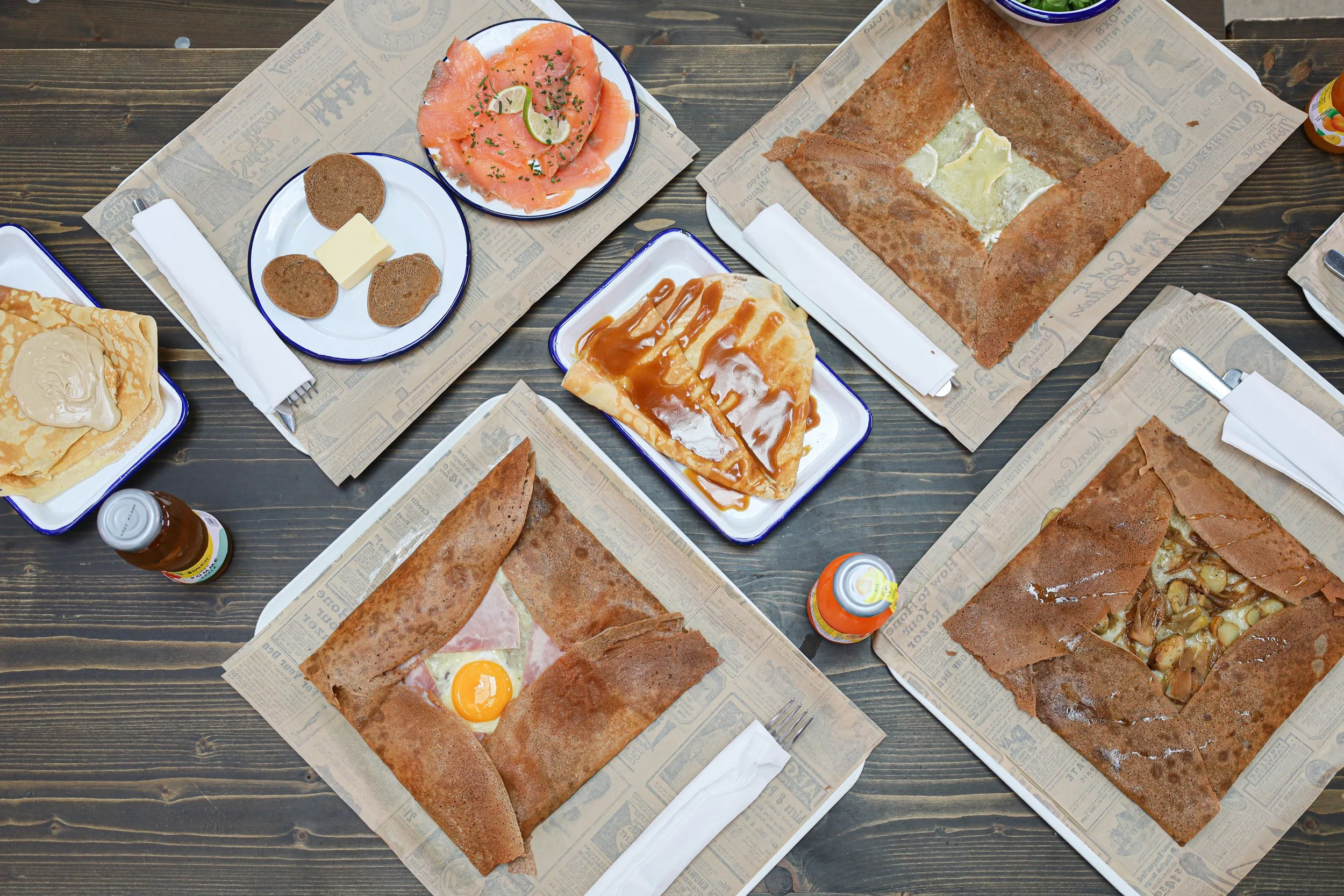 Petit déjeuner avec crêpes, pancakes, saumon fumé, viande hachée, œuf au plat, galette bretonne, et boissons sur une table en bois.