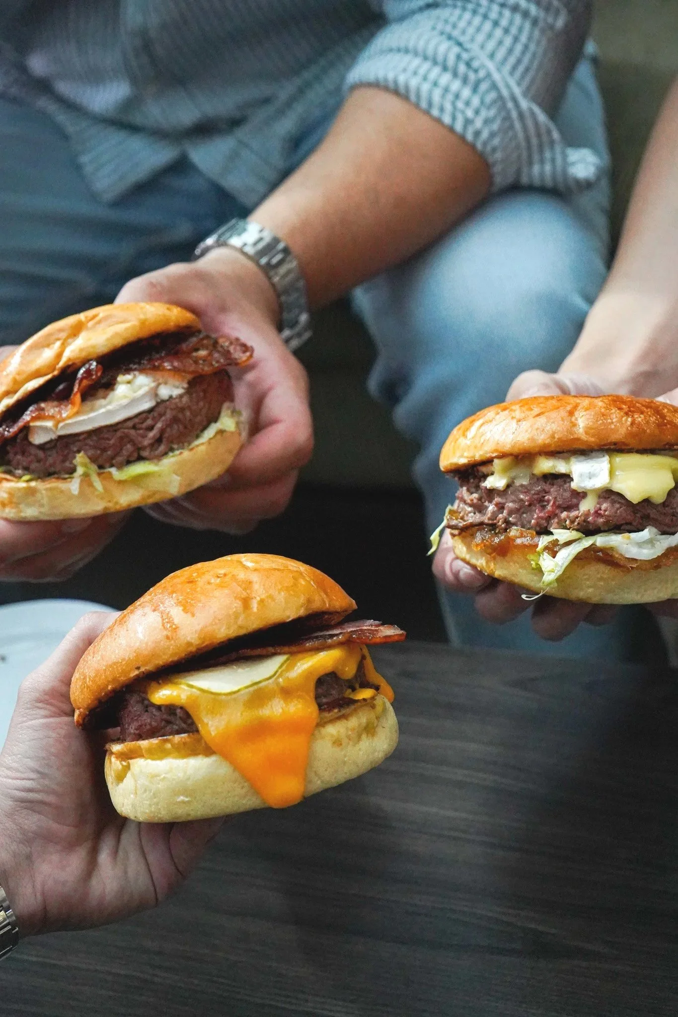 Trois mains tenant des burger avec du fromage, de la viande, de la laitue et du bacon, sur une table en bois.