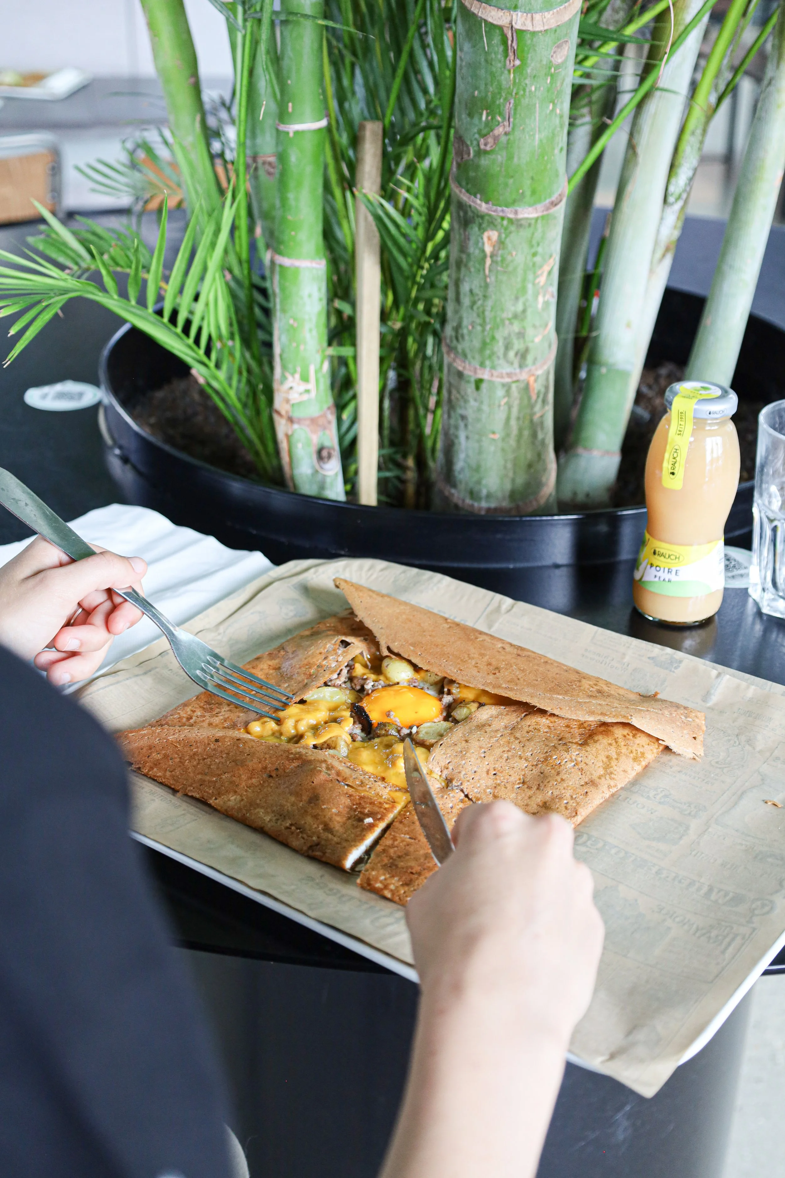 Une personne coupe une galette de sarrasin fourrée, probablement une crêpe bretonne, avec un œuf au centre, sur une assiette recouverte de papier parchemin, près d'une plante verte en pot, dans un lieu intérieur.