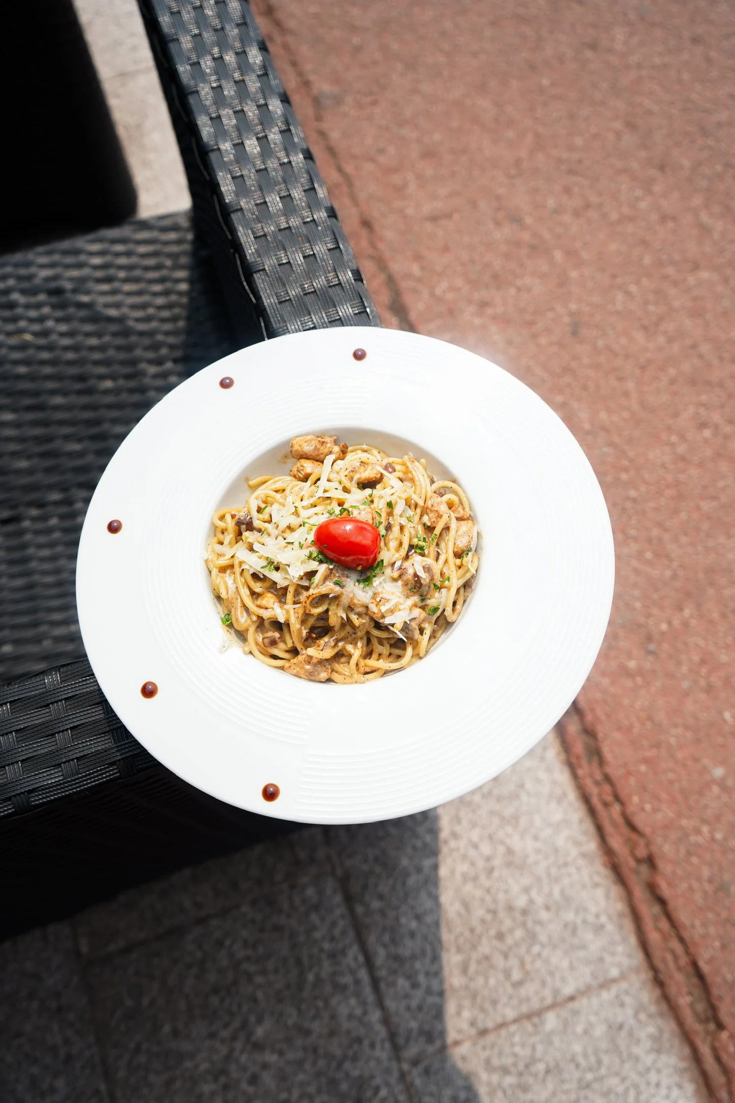Assiette de sauce carbonara avec des pâtes, une tomate cerise rouge, du parmesan râpé, et une garniture de persil, posée sur une table extérieure en osier noir.
