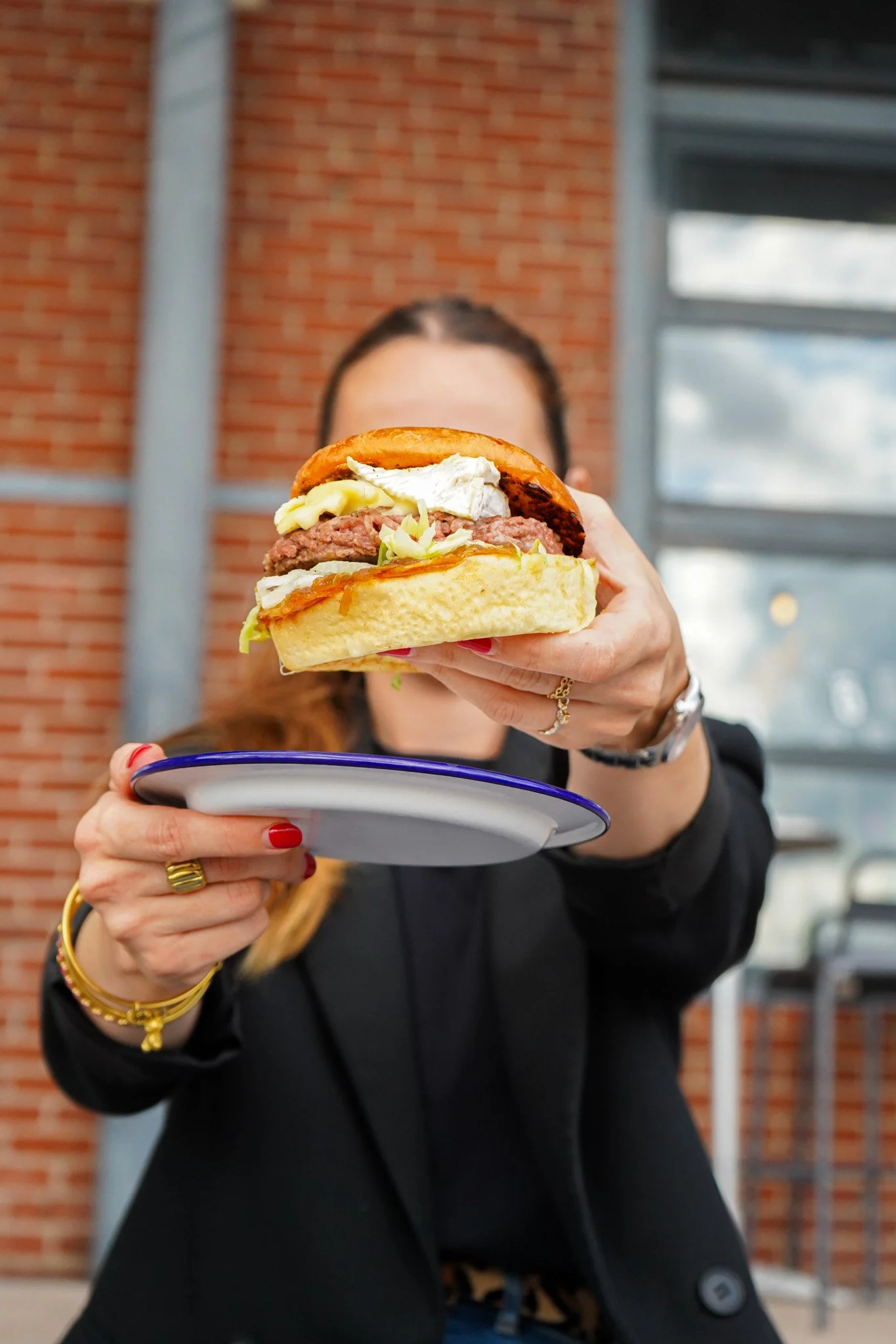 Une femme tient un burger avec du pain, une tranche de viande, de la salade, du fromage et une sauce, devant un mur en briques.