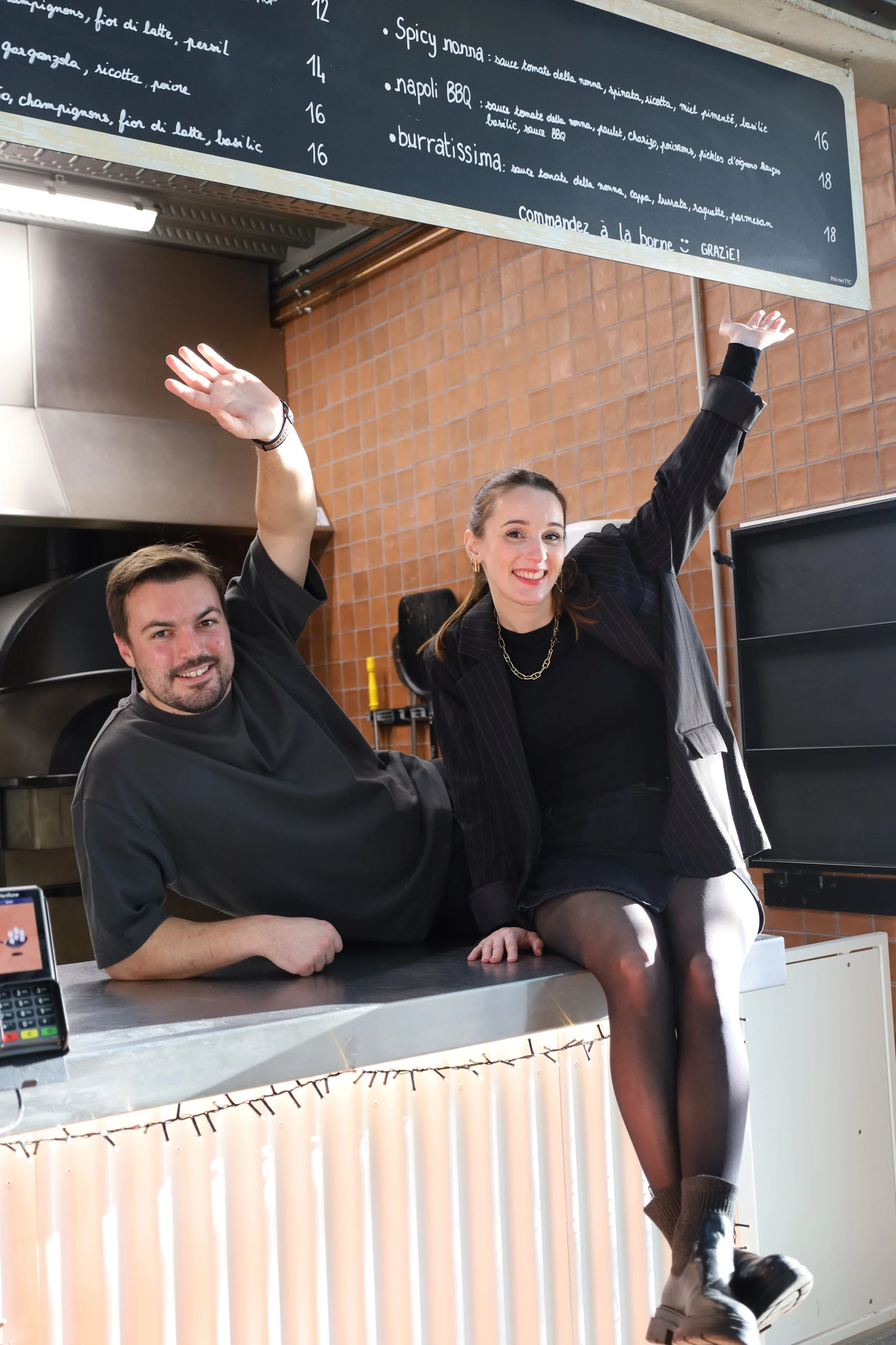 Deux personnes souriantes et joyeuses posent ensemble derrière un comptoir, la femme est assise sur le comptoir avec une jambe croisée, portant une veste noire, un t-shirt noir, une jupe noire, des collants et des bottes, tandis que l'homme est debout à côté d'elle. Un menu en français est visible au-dessus d'eux.