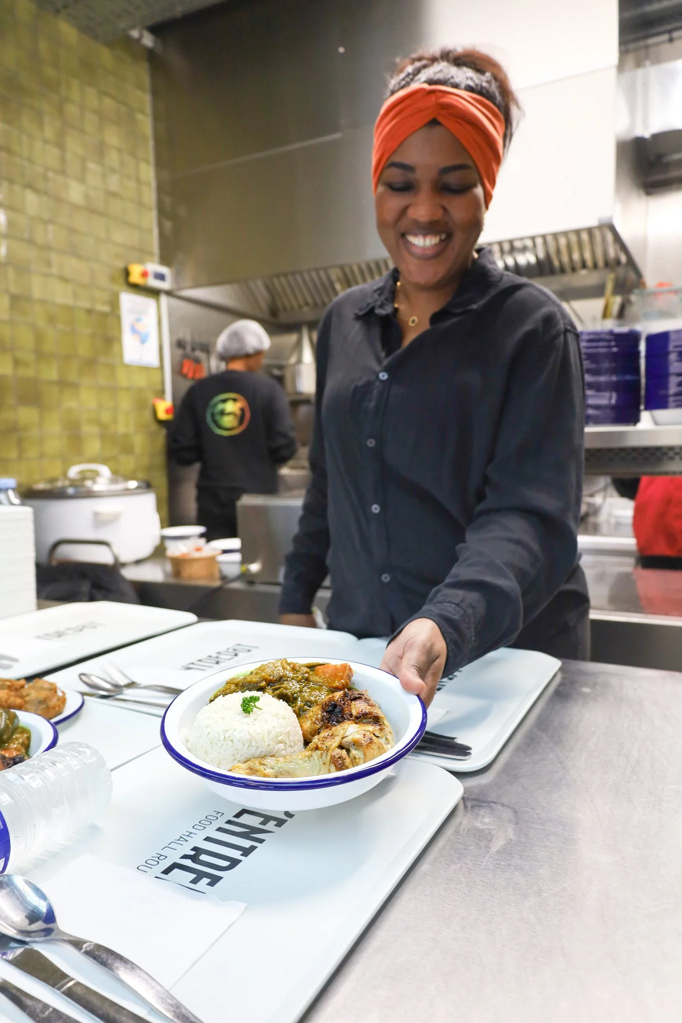 Une femme souriante présente un plat avec du riz, de la viande et un curry, dans une cuisine de restaurant.