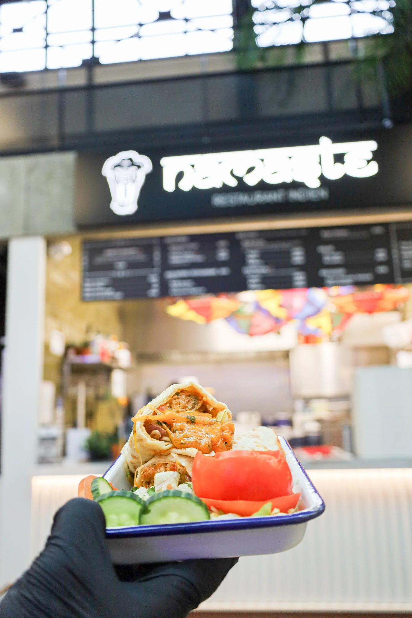Une main déguisée avec un gant noir tenant une assiette contenant un rouleau de printemps, des tranches de concombre, une tranche de tomate et de la laitue, devant un stand de restauration rapide nommé 'Pizzeria'.