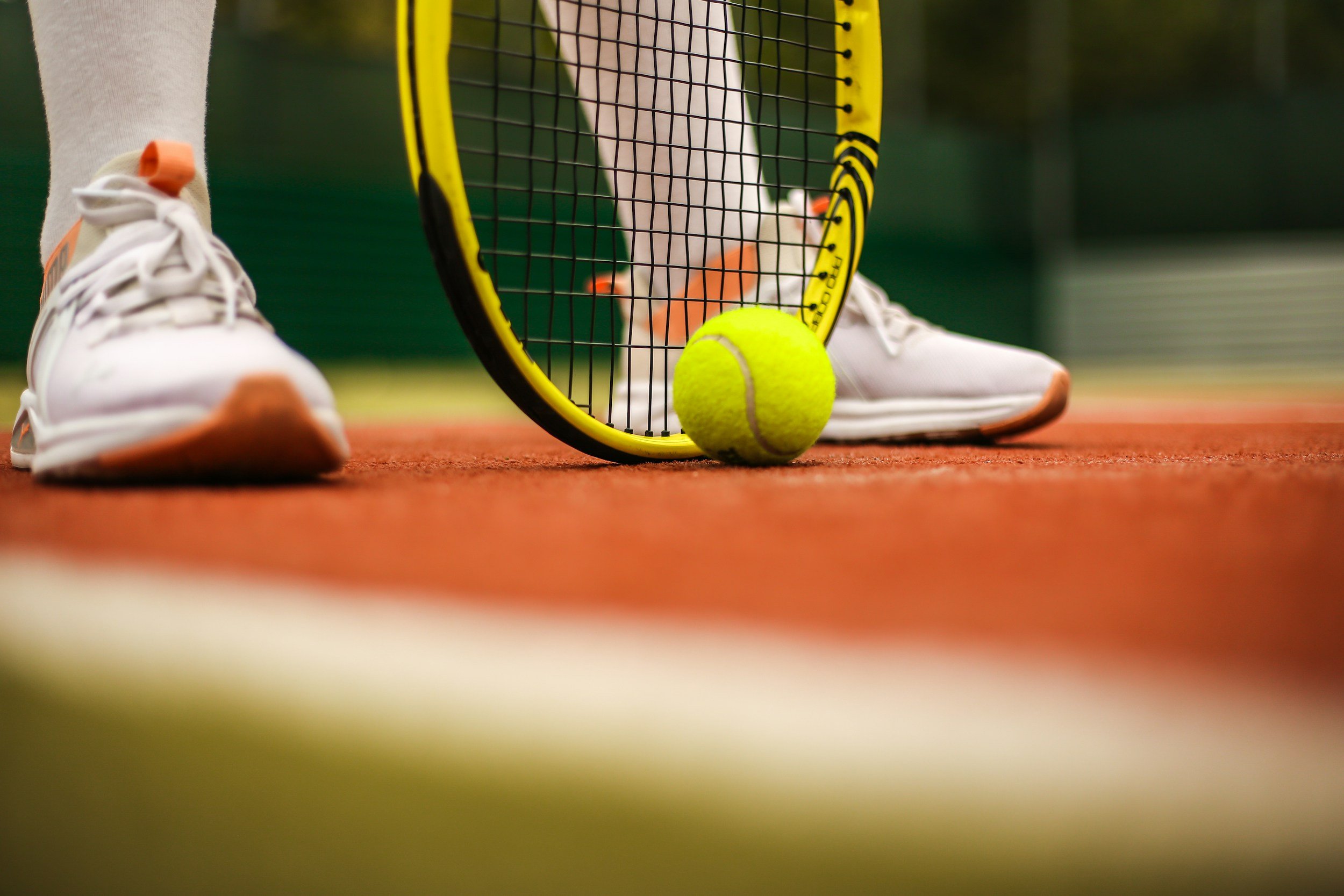 Close-up of a tennis court with a tennis ball, tennis racket, and a person's feet wearing white shoes and gray pants.