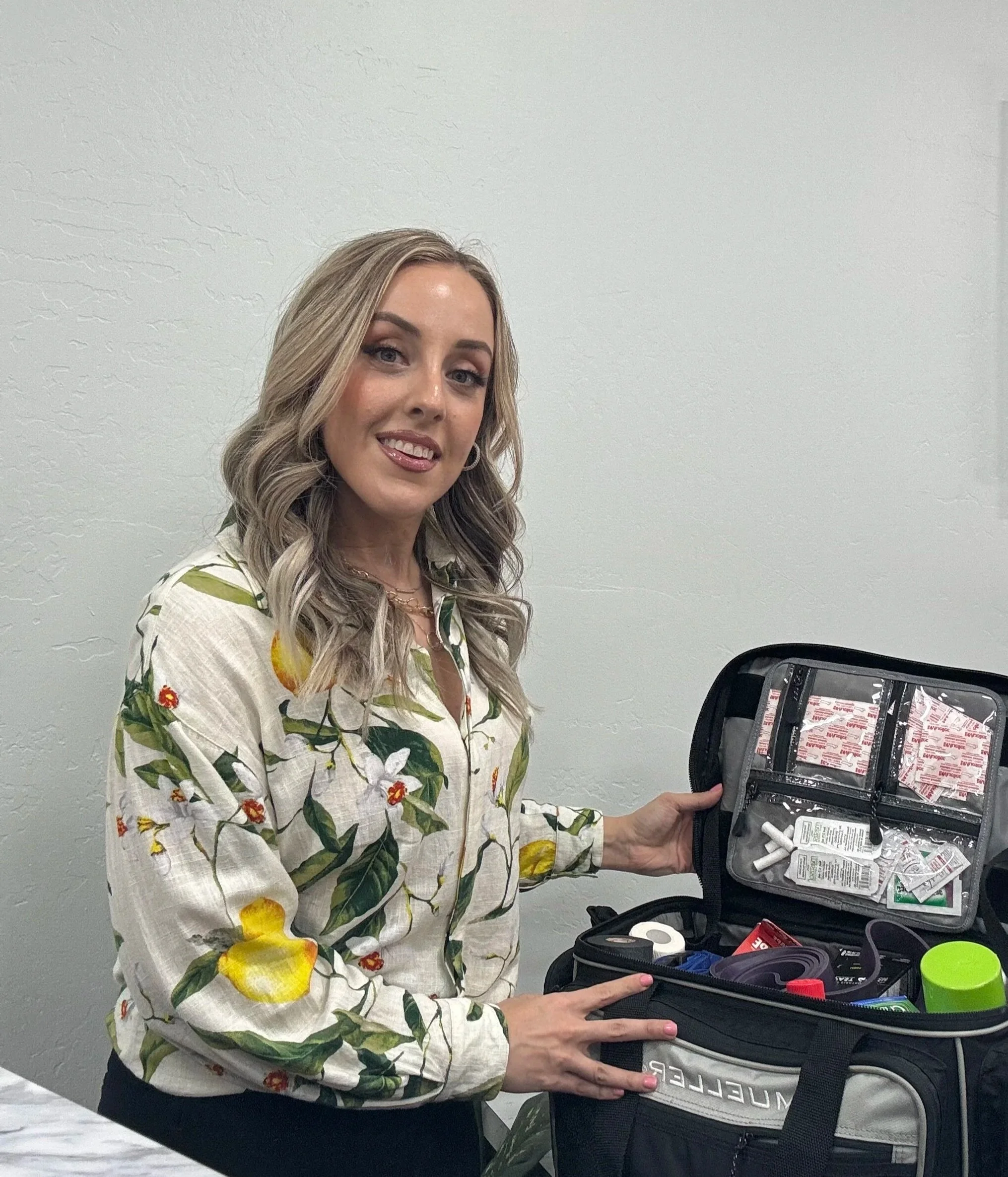 A woman with long, wavy, blonde hair wearing a floral blouse standing next to an open first aid kit.