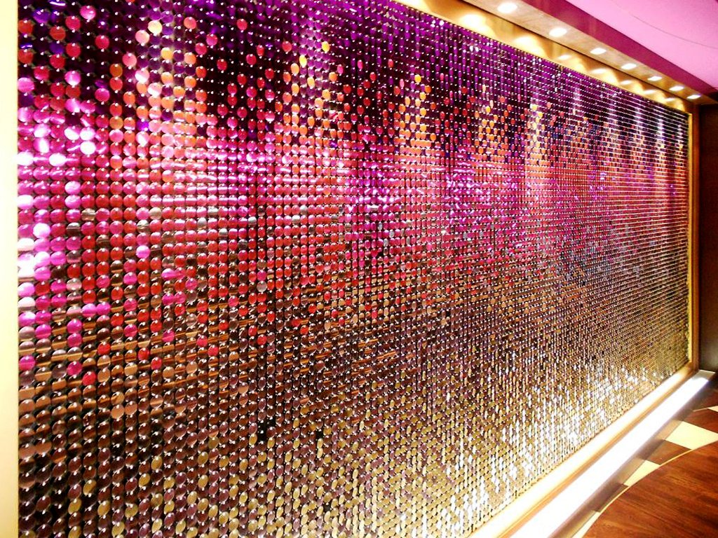 Bunt schimmernde, reflektierende Pailletten auf einer Wand in Pink, Gold und Silber Tönen, mit Beleuchtung an der Oberseite.