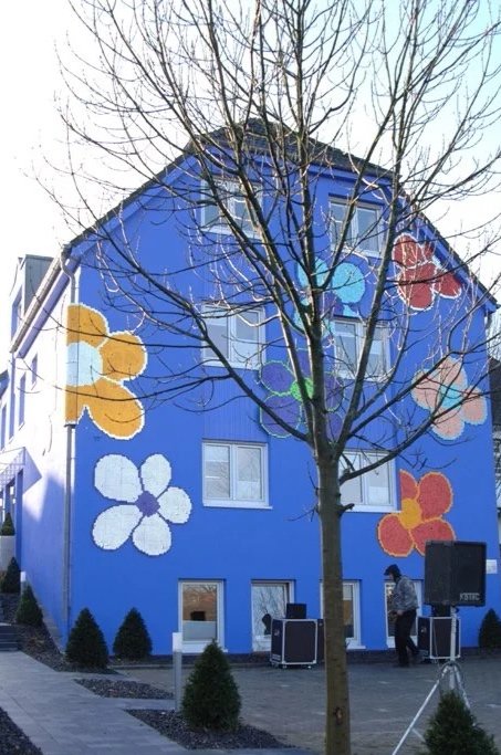 Bunt bemalte blumenartige Muster an einem blauen Haus, im Vordergrund ein Baum und eine Person, die vor dem Haus steht.