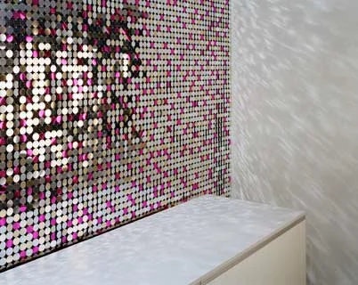 Eine Wand aus kleinen, runden, bunten Steinen in Schwarz, Weiß und Pink, gegenüber einer weißen Wand, mit einer weißen Kommode davor.