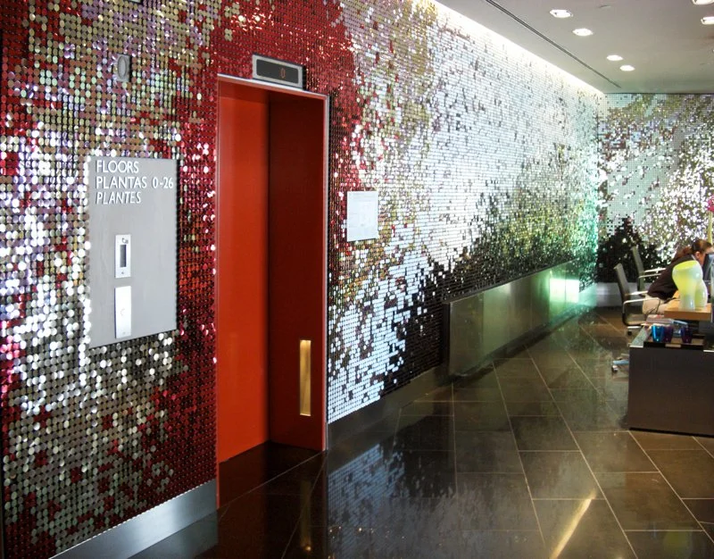 Aufzug mit esculthartigem, spiegelndem Wanddesign im Hintergrund, Sitzbereich mit Personen im rechten Bereich, schwarze Fliesenboden.