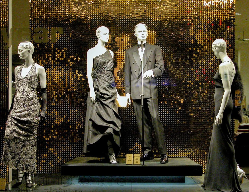 Modell-Displays in einem Schaufenster mit eleganter Abendgarderobe, bestehend aus fünf weißen Mannequins, die schwarze und goldene Kleider und Anzüge tragen, vor einem glitzernden goldenen Hintergrund.