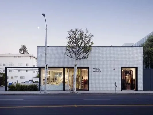 Straßenszene mit einem modernen Gebäude, einem Baum in der Mitte und einem beleuchteten Laden im Vordergrund.