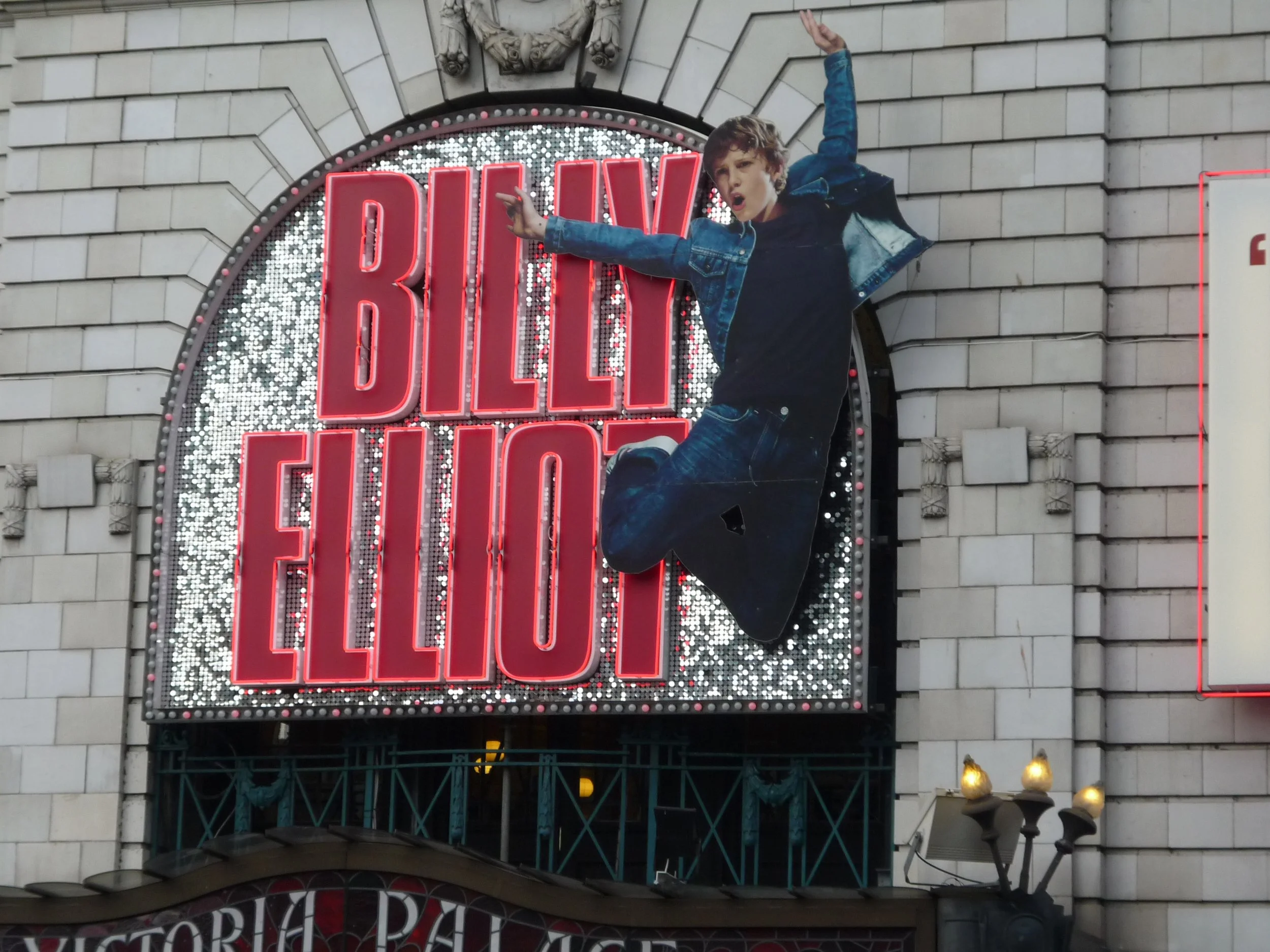 Werbetafel mit dem Schriftzug 'BILLY ELLIOT' in roten Buchstaben, darüber eine lebensgroße Statue eines Jungen in Jeansjacke, die springt.