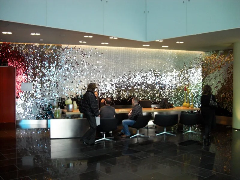 Menschen sitzen und stehen in einem modernen Empfangsbereich mit einer auffälligen, mosaikartigen Wand hinter dem Empfangstresen.