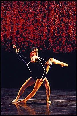 Zwei Tänzer in schwarzen Kostümen auf der Bühne, einer hebt den anderen in einer tänzerischen Pose, im Hintergrund rote Lichter.