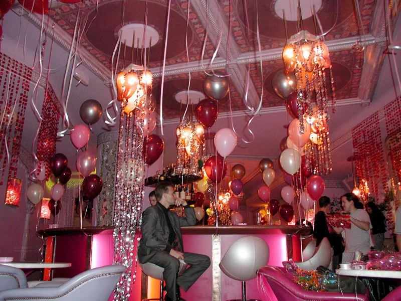 Innenraum einer Party mit vielen bunten Ballons, Lichter und festlicher Dekoration, Gäste feiern an der Bar.