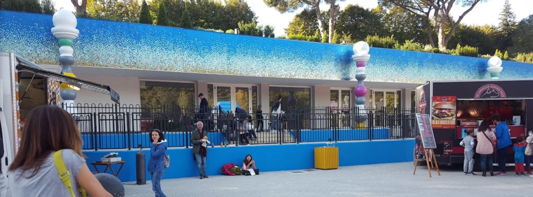Ein Freizeitpark mit einem bunten, blauen Gebäude, das eine obere Terrasse hat, auf der einige Menschen sitzen und spazieren gehen. Es gibt Food-Trucks, eine Gruppe von Leuten, die am Stand Schlange stehen, und im Vordergrund eine Frau mit braunen Haaren und einer gelben Tasche.
