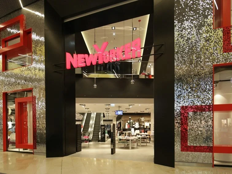 Eingangsbereich eines Einzelhandelsgeschäfts mit einem leuchtenden rosa NY-Filiale-Schild und einer goldenen, funkelnden Wanddekoration.