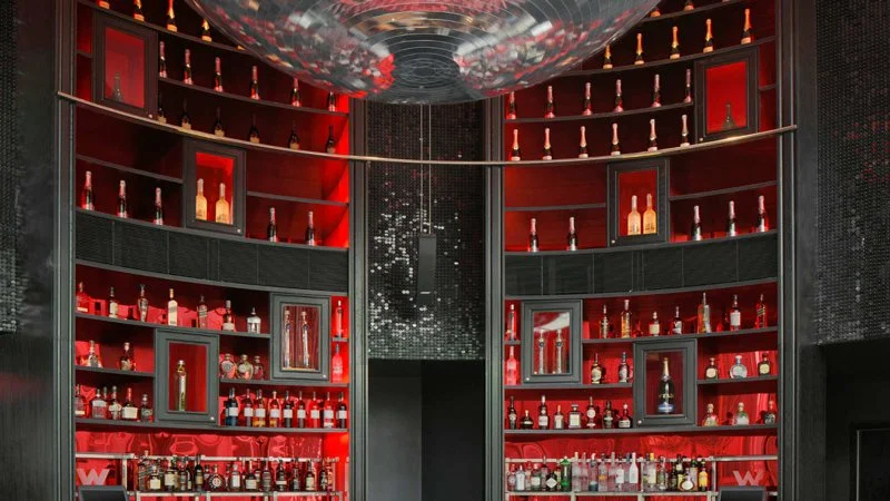 Ein moderner Barbereich mit roten Regalen, die verschiedene Spirituosenflaschen präsentieren, und einer dunklen, eleganten Atmosphäre.