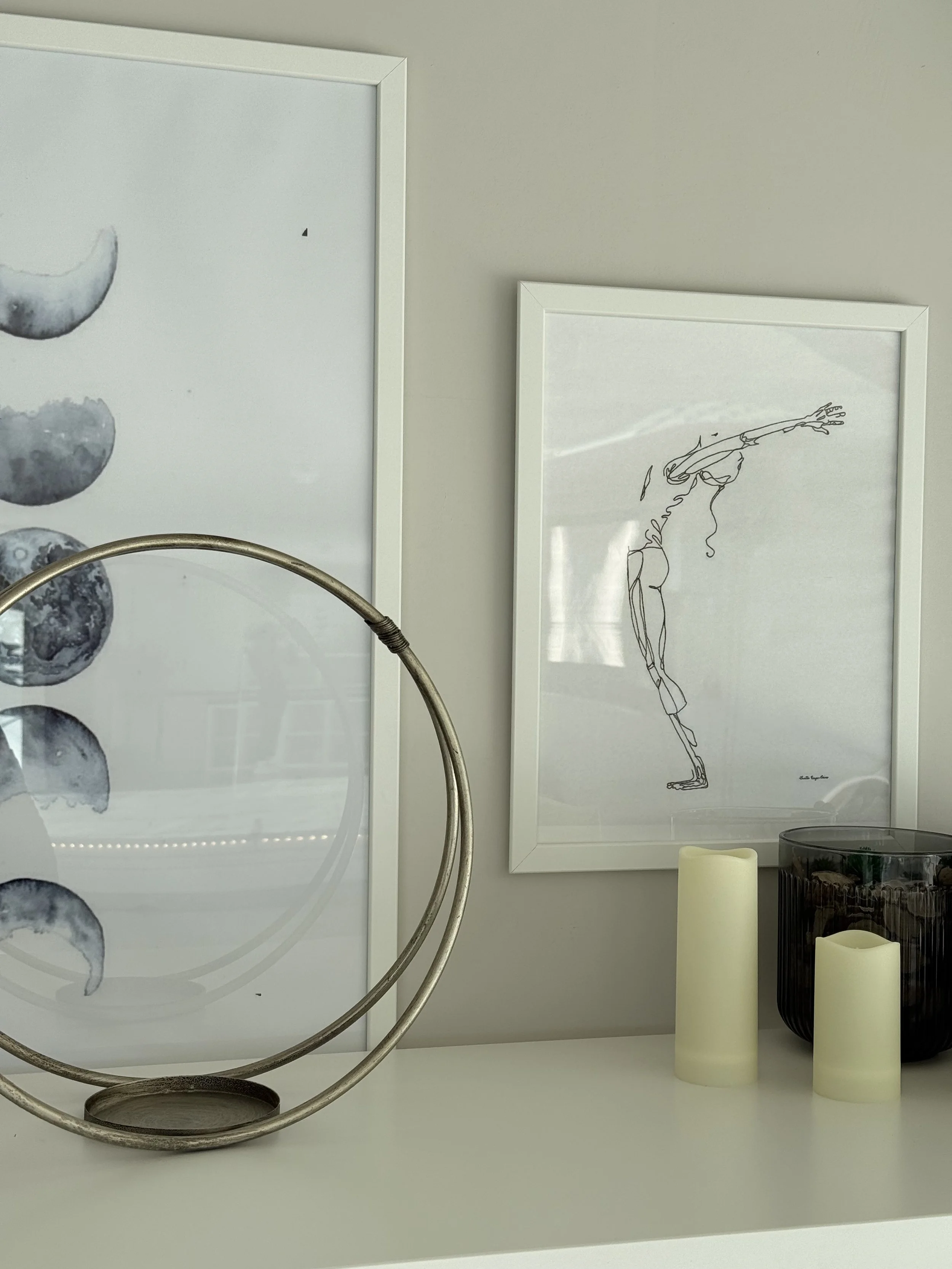 Decorative interior with two framed black-and-white drawings, one of a moon phases chart and the other of a woman in a stretching pose, along with white candles and a black vase on a white surface.