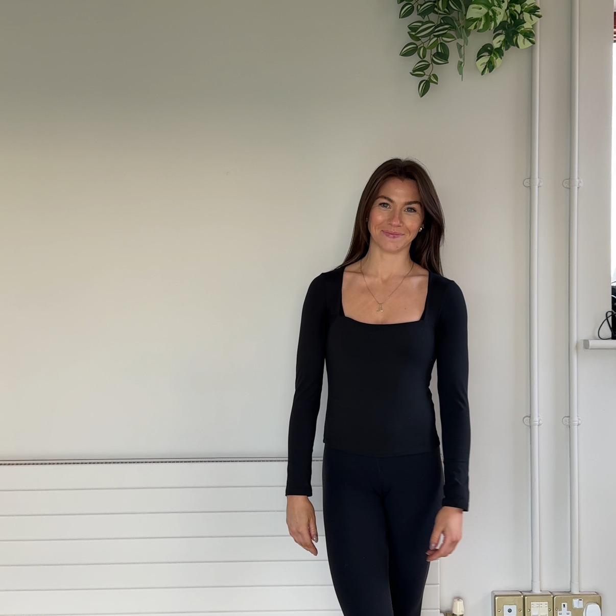 A woman with long brown hair wearing a black top and pants standing indoors against a plain light wall with some greenery and electrical wiring in the background.