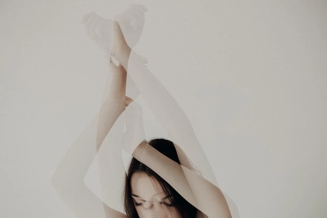 A woman with dark hair has her eyes closed and is lifting her arms above her head, creating a layered, abstract effect with her arms and face against a plain background.