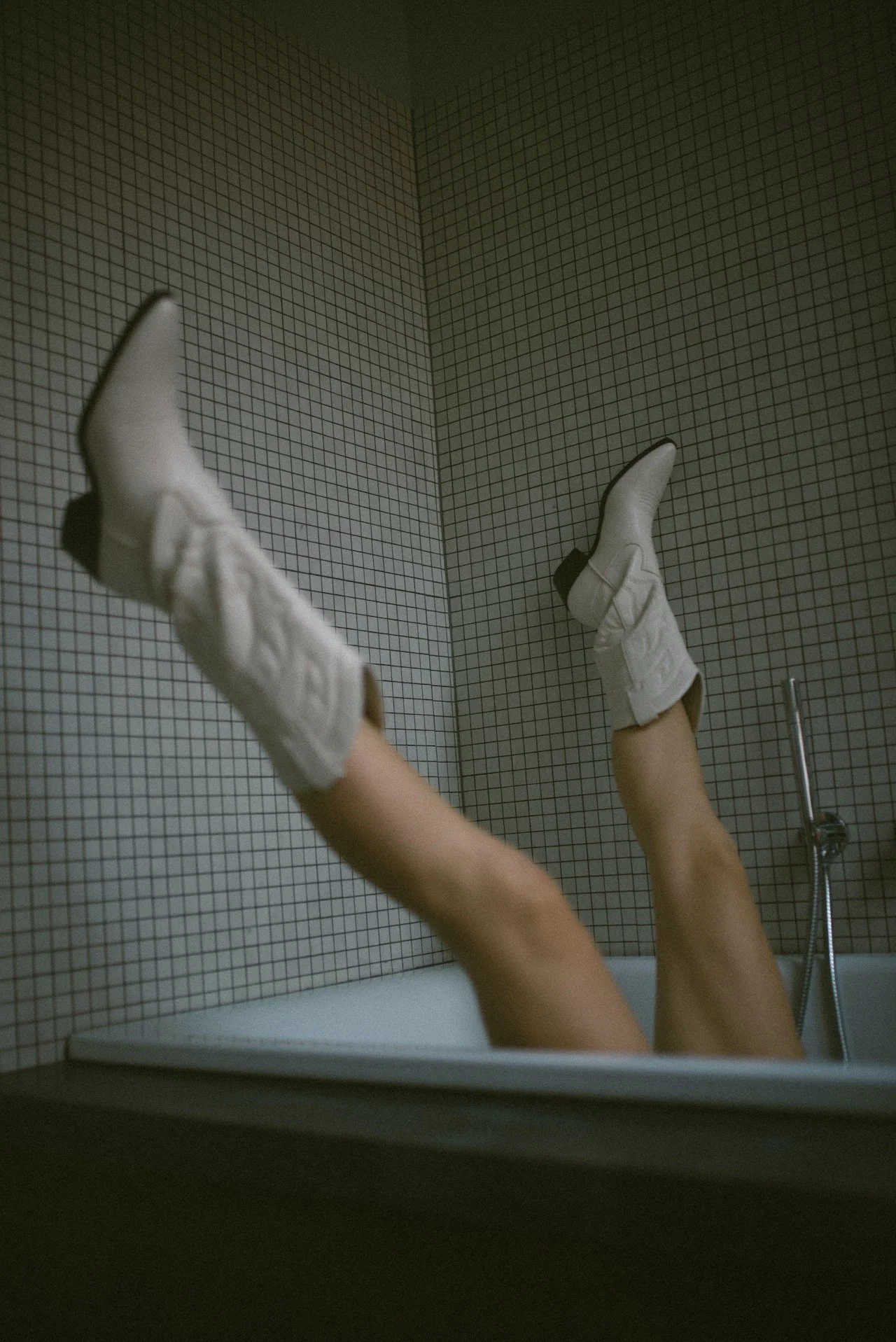 A person lying in a bathtub with legs raised, wearing white boots and beige skin. The bathtub is against a wall covered with small brown tiles, and a shower fixture is visible on the right.